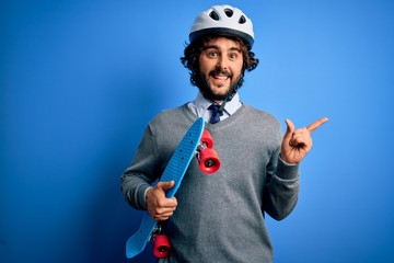 Handsome skater man with beard wearing security helmet holding skate over blue background very happy pointing with hand and finger to the side
