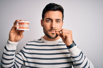 Young handsome man with beard holding plastic denture teeth over white background mouth and lips shut as zip with fingers. Secret and silent, taboo talking