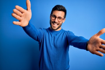 Young handsome man with beard wearing casual sweater and glasses over blue background looking at...