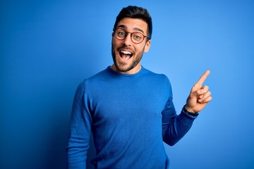 Young handsome man with beard wearing casual sweater and glasses over blue background with a big...