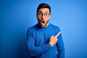 Young handsome man with beard wearing casual sweater and glasses over blue background Surprised pointing with finger to the side, open mouth amazed expression. - obrazy, fototapety, plakaty