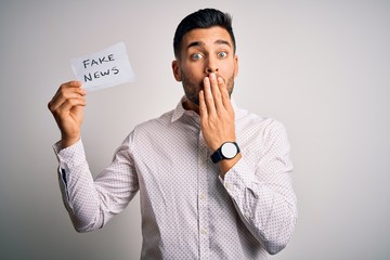 Young business man holding fake news paper over isolated background cover mouth with hand shocked...
