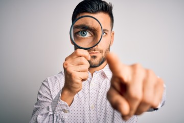 Young detective man looking through magnifying glass over isolated background pointing with finger to the camera and to you, hand sign, positive and confident gesture from the front