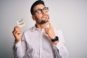 Young handsome man holding reminder paper with pay taxes message over white background serious face...