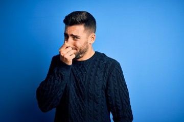 Young handsome man wearing casual sweater standing over isolated blue background smelling something...