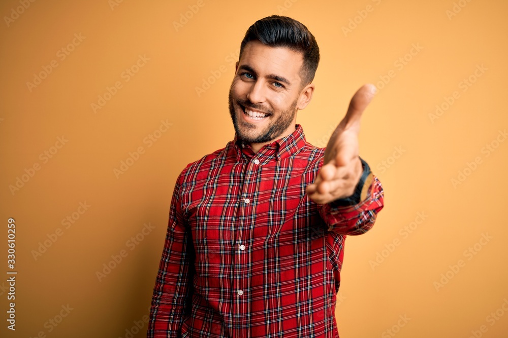 Poster Young handsome man wearing casual shirt standing over isolated yellow background smiling friendly offering handshake as greeting and welcoming. Successful business.