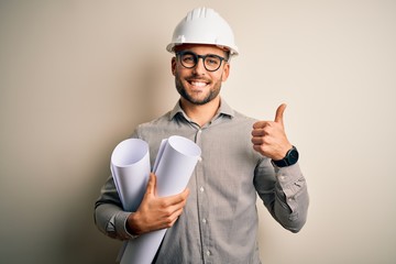 Young architect man wearing contractor helmet holding project paper plan over isolated background happy with big smile doing ok sign, thumb up with fingers, excellent sign - obrazy, fototapety, plakaty