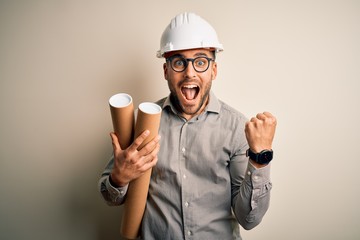 Young architect man wearing contractor helmet holding project paper plan over isolated background screaming proud and celebrating victory and success very excited, cheering emotion