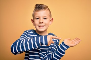 Young little caucasian kid with blue eyes wearing nautical striped shirt over yellow background amazed and smiling to the camera while presenting with hand and pointing with finger.