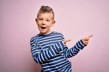 Young little caucasian kid with blue eyes wearing nautical striped shirt over pink background smiling and looking at the camera pointing with two hands and fingers to the side.
