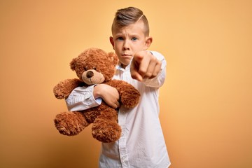 Young little caucasian kid hugging teddy bear stuffed animal over yellow background pointing with...