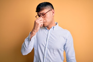 Young handsome hispanic business man wearing nerd glasses over yellow background smelling something...