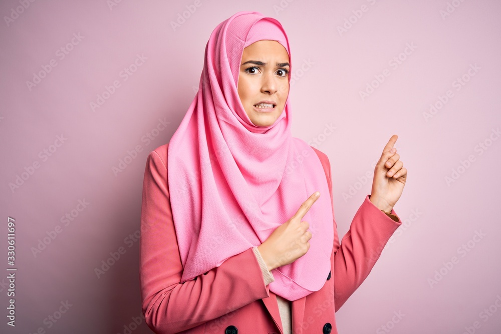 Sticker young beautiful girl wearing muslim hijab standing over isolated pink background pointing aside worr