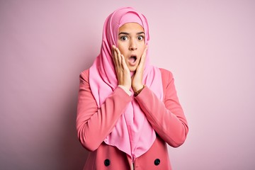 Young beautiful girl wearing muslim hijab standing over isolated pink background afraid and shocked, surprise and amazed expression with hands on face