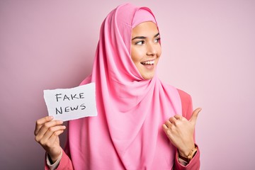 Young beautiful girl wearing muslim hijab holding paper with fake news message pointing and showing with thumb up to the side with happy face smiling