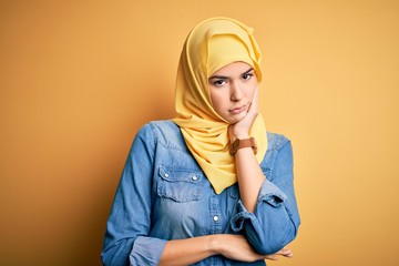Young beautiful girl wearing muslim hijab standing over isolated yellow background thinking looking...