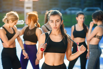 Pretty young woman with jump rope on her neck looking at camera after outdoors workout with her female friends on the background on the stadium.