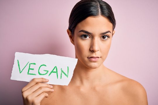 Young beautiful girl holding paper with vegan message over isolated pink background with a confident expression on smart face thinking serious