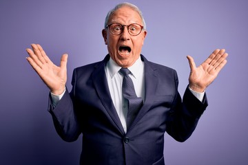 Grey haired senior business man wearing glasses and elegant suit and tie over purple background celebrating crazy and amazed for success with arms raised and open eyes screaming excited. Winner