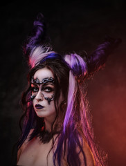 Portrait of a demonic girl wearing scary fantasy make up and violet horns posing in a dark studio on a grey background looking scary