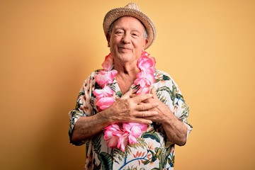 Grey haired senior man wearing summer hat and hawaiian lei over yellow background smiling with...