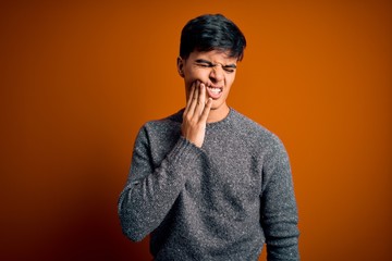 Young handsome man wearing casual sweater standing over isolated orange background touching mouth with hand with painful expression because of toothache or dental illness on teeth. Dentist