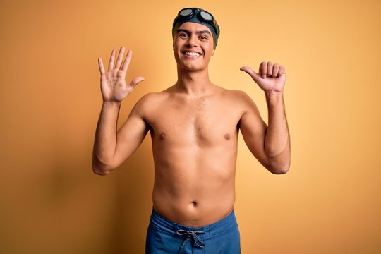 Young handsome man shirtless wearing swimsuit and swim cap over isolated yellow background showing and pointing up with fingers number six while smiling confident and happy.