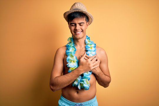 Young handsome tourist man on vacation wearing swimwear and hawaiian lei flowers smiling with hands on chest with closed eyes and grateful gesture on face. Health concept.