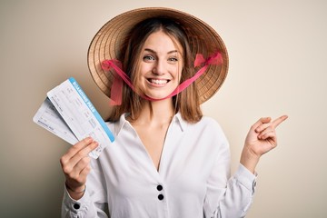 Young beautiful redhead tourist woman wearing asian traditional hat holding boarding pass very happy pointing with hand and finger to the side