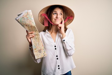 Young beautiful redhead woman wearing asian traditional hat holding city map serious face thinking about question, very confused idea