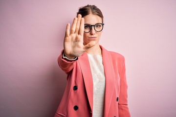 Young beautiful redhead woman wearing jacket and glasses over isolated pink background doing stop...