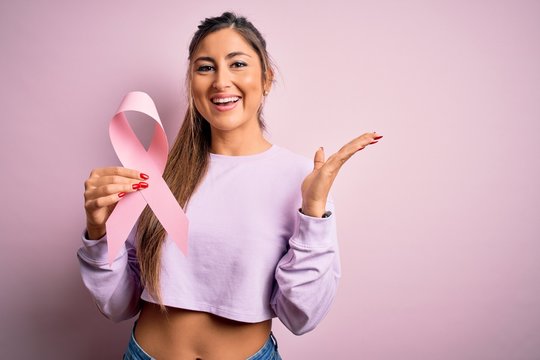 Young beautiful brunette woman holding pink cancer ribbon symbol over isolated background very happy and excited, winner expression celebrating victory screaming with big smile and raised hands