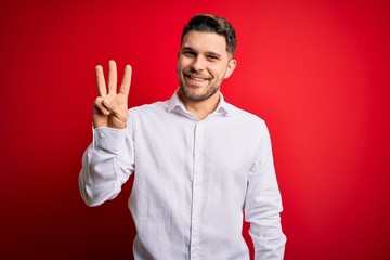 Young business man with blue eyes wearing elegant shirt standing over red isolated background showing and pointing up with fingers number three while smiling confident and happy.