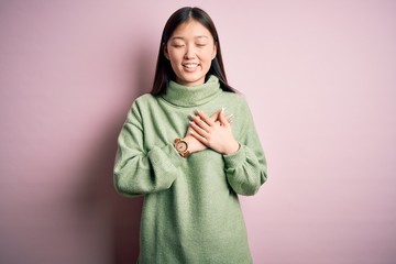 Young beautiful asian woman wearing green winter sweater over pink solated background smiling with hands on chest with closed eyes and grateful gesture on face. Health concept.