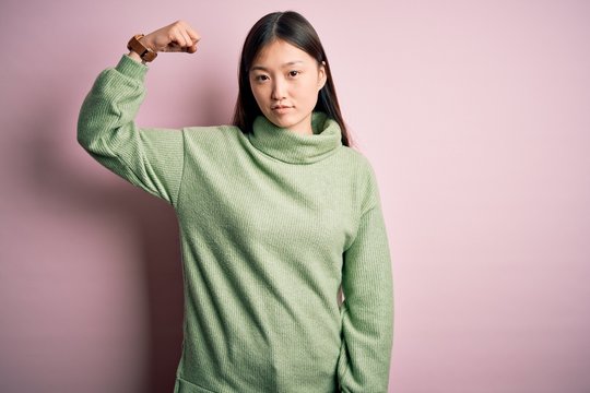 Young beautiful asian woman wearing green winter sweater over pink solated background Strong person showing arm muscle, confident and proud of power