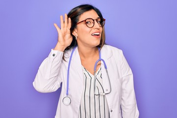 Professional doctor woman wearing stethoscope and medical coat over purple background smiling with hand over ear listening an hearing to rumor or gossip. Deafness concept.