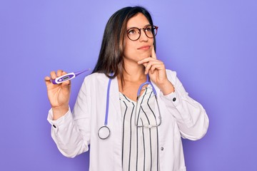 Professional doctor woman showing medical thermometer over purple background serious face thinking...