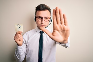 Young call center operator man wearing headset holding paper note with question mark with open hand doing stop sign with serious and confident expression, defense gesture