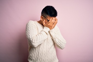 Young handsome latin man wearing white casual sweater and glasses over pink background with sad expression covering face with hands while crying. Depression concept.