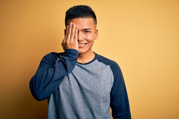 Young handsome latin man wearing casual t-shirt standing over yellow background covering one eye with hand, confident smile on face and surprise emotion.