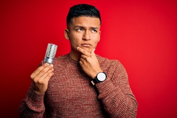 Young handsome latin man holding led lightbulb over isolated red background serious face thinking...