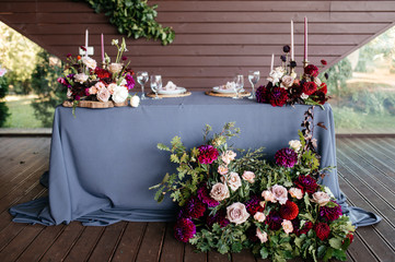 Fototapeta na wymiar The festive table for the bride and groom is decorated with a blue tablecloth with beautiful floral arrangements of different blossom flowers and greenery on the table and near the table and candles