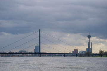 View of city on riverside, Rhine Tower, suspension bridge and Rhine River with beautiful dramatic cloudy and overcast golden sky during twilight sunset time after storm.