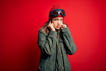 Young blonde girl wearing ski glasses and winter coat for ski weather over red background covering ears with fingers with annoyed expression for the noise of loud music. Deaf concept.