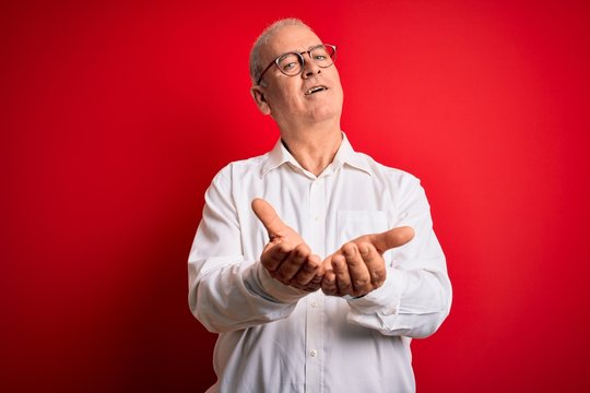 Middle age handsome hoary man wearing casual shirt and glasses over red background Smiling with hands palms together receiving or giving gesture. Hold and protection