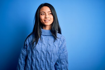 Young beautiful chinese woman wearing casual sweater over isolated blue background looking away to side with smile on face, natural expression. Laughing confident.