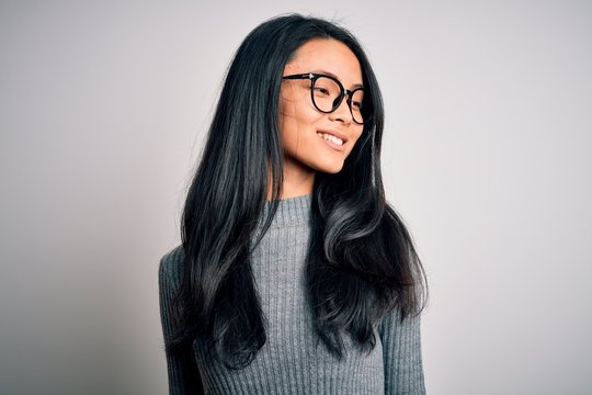 Young beautiful chinese woman wearing glasses and sweater over isolated white background looking away to side with smile on face, natural expression. Laughing confident.