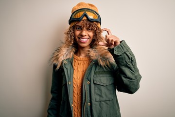 Young african american skier woman with curly hair wearing snow sportswear and ski goggles smiling and confident gesturing with hand doing small size sign with fingers looking and the camera. Measure