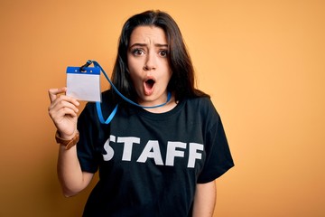 Young brunette worker woman wearing staff t-shirt as uniform showing id card scared in shock with a...