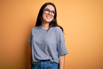 Young brunette woman wearing glasses and navy t-shirt over yellow isolated background looking away to side with smile on face, natural expression. Laughing confident.